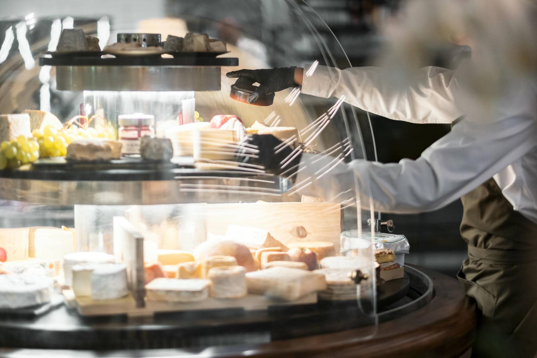 un homme portant des gants noirs se tient derrière une vitrine remplie de fromages