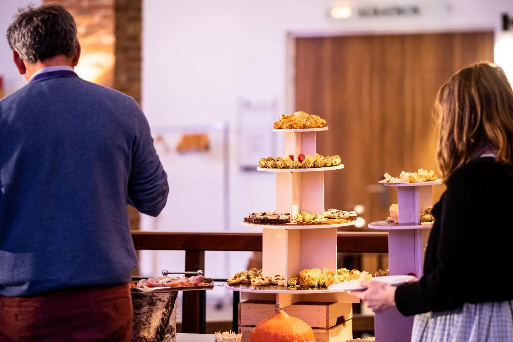 gros plan sur deux personnes se servant à un cocktail dînatoire dressé en salle de réunion, autour d’une offre salée composée de fromages et de charcuterie