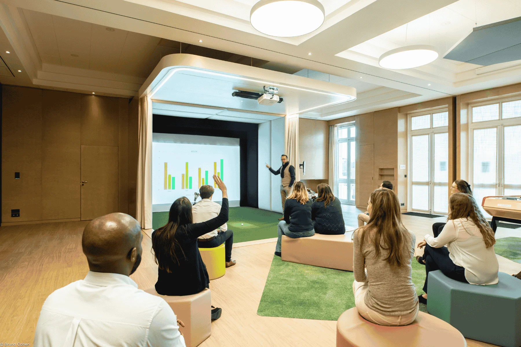 salle de réunion aux tons pastel, équipée d’un écran de projection mural, avec des participants face à un intervenant, un accès direct vers le golf et des assises informelles composées de poufs colorés