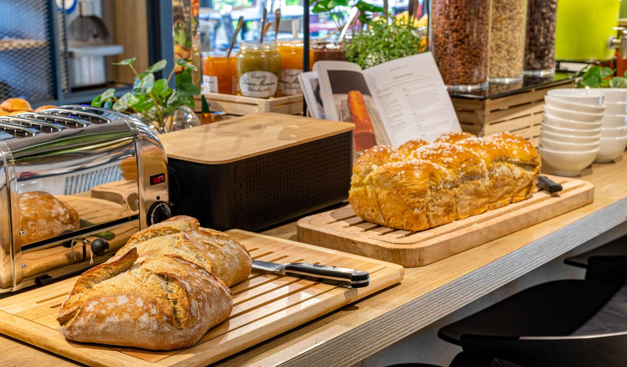 Comptoir de buffet petit-déjeuner d'hôtel proposant de la restauration sur place avec un plan de travail en bois clair, deux pains de campagne croustillants et une brioche sucrée sur des planches en bois au premier plan, un grille-pain en inox et une boîte à pain noire à gauche, des pots de confiture et de miel avec des distributeurs de céréales en verre et des plantes vertes en arrière-plan.
