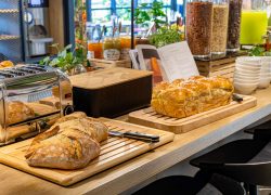 Comptoir de buffet petit-déjeuner d'hôtel proposant de la restauration sur place avec un plan de travail en bois clair, deux pains de campagne croustillants et une brioche sucrée sur des planches en bois au premier plan, un grille-pain en inox et une boîte à pain noire à gauche, des pots de confiture et de miel avec des distributeurs de céréales en verre et des plantes vertes en arrière-plan.