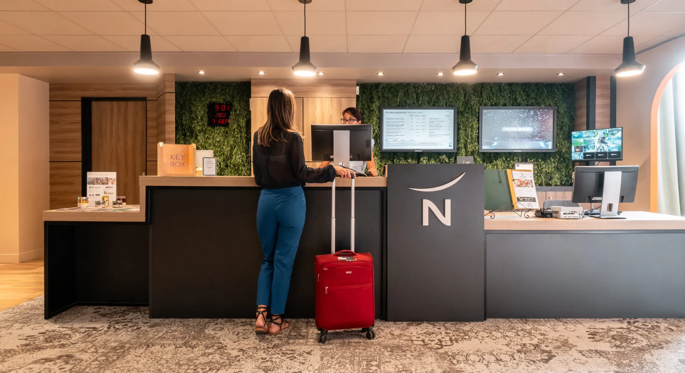 une femme avec une valise rouge se tient devant un comptoir sur lequel figure le logo Novotel