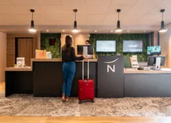 une femme avec une valise rouge se tient devant un comptoir sur lequel figure le logo Novotel