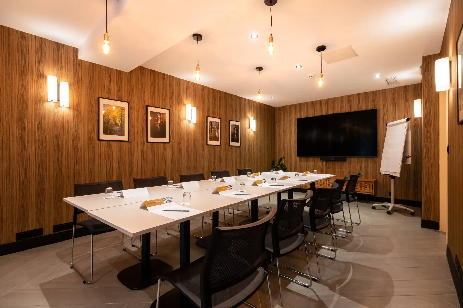 en::Meeting room arranged in boardroom style with tables facing each other, chairs on both sides, wood-effect wall, large screen, hanging light bulbs, and wall lights. fr::Salle de réunion avec tables disposées en configuration board, chaises de chaque côté, mur effet bois, grand écran, ampoules suspendues au plafond et luminaires muraux.
