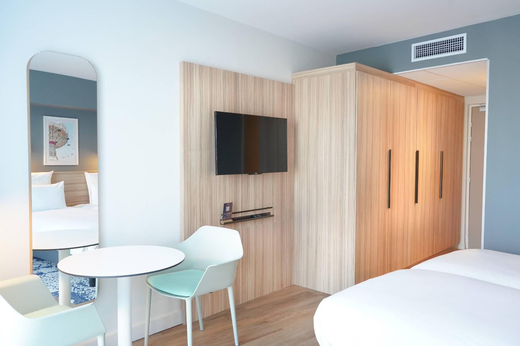 en::White table paired with a light blue chair, wooden unit integrating a wardrobe and TV stand, with the corner of the bed visible. fr::Table blanche accompagnée d’une chaise bleu clair, meuble en bois intégrant une armoire et un support TV, avec le coin du lit visible.