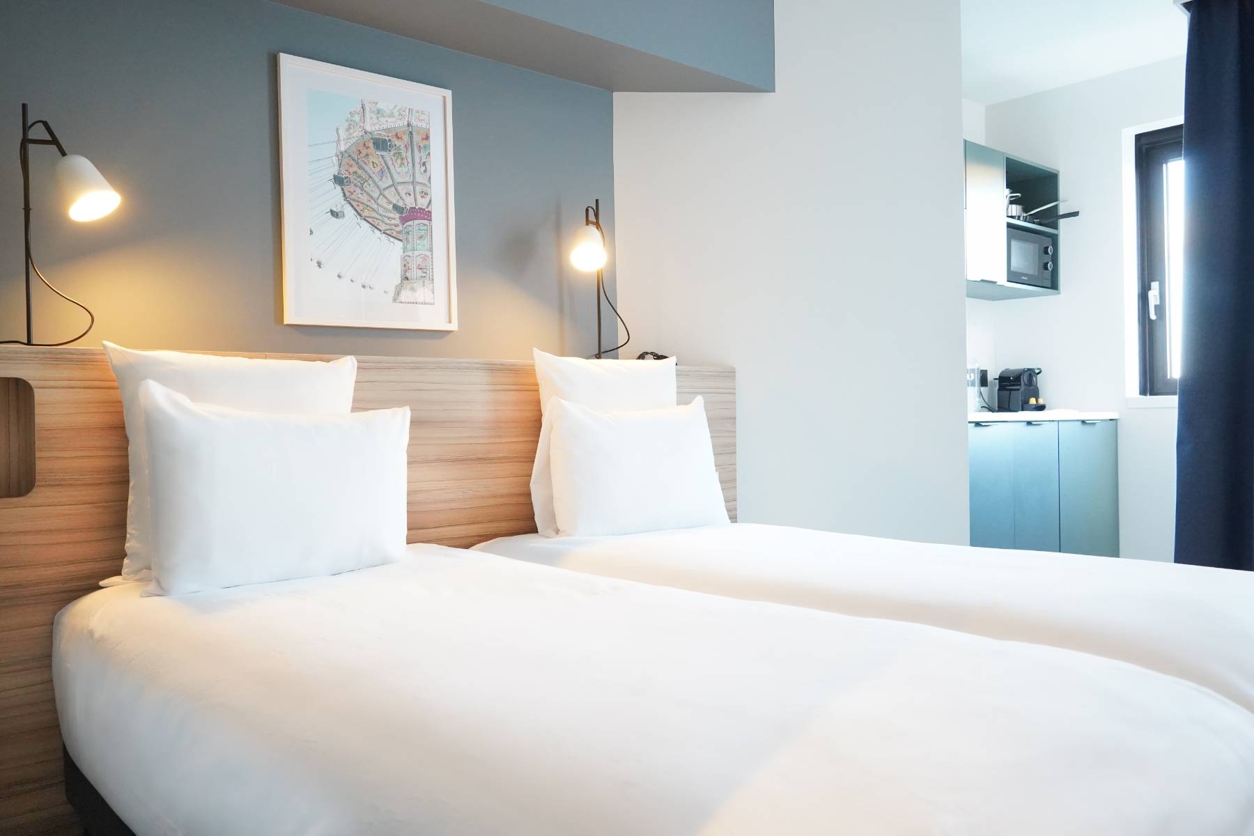 en::Twin room with a wooden headboard, grey and white wall, bedside lights on, a fully equipped royal blue kitchen, separated by a thick navy curtain. fr::Chambre avec lits jumeaux et tête de lit en bois, mur gris et blanc, lampes de chevet allumées, cuisine équipée bleu roi, séparée par un rideau épais bleu marine.