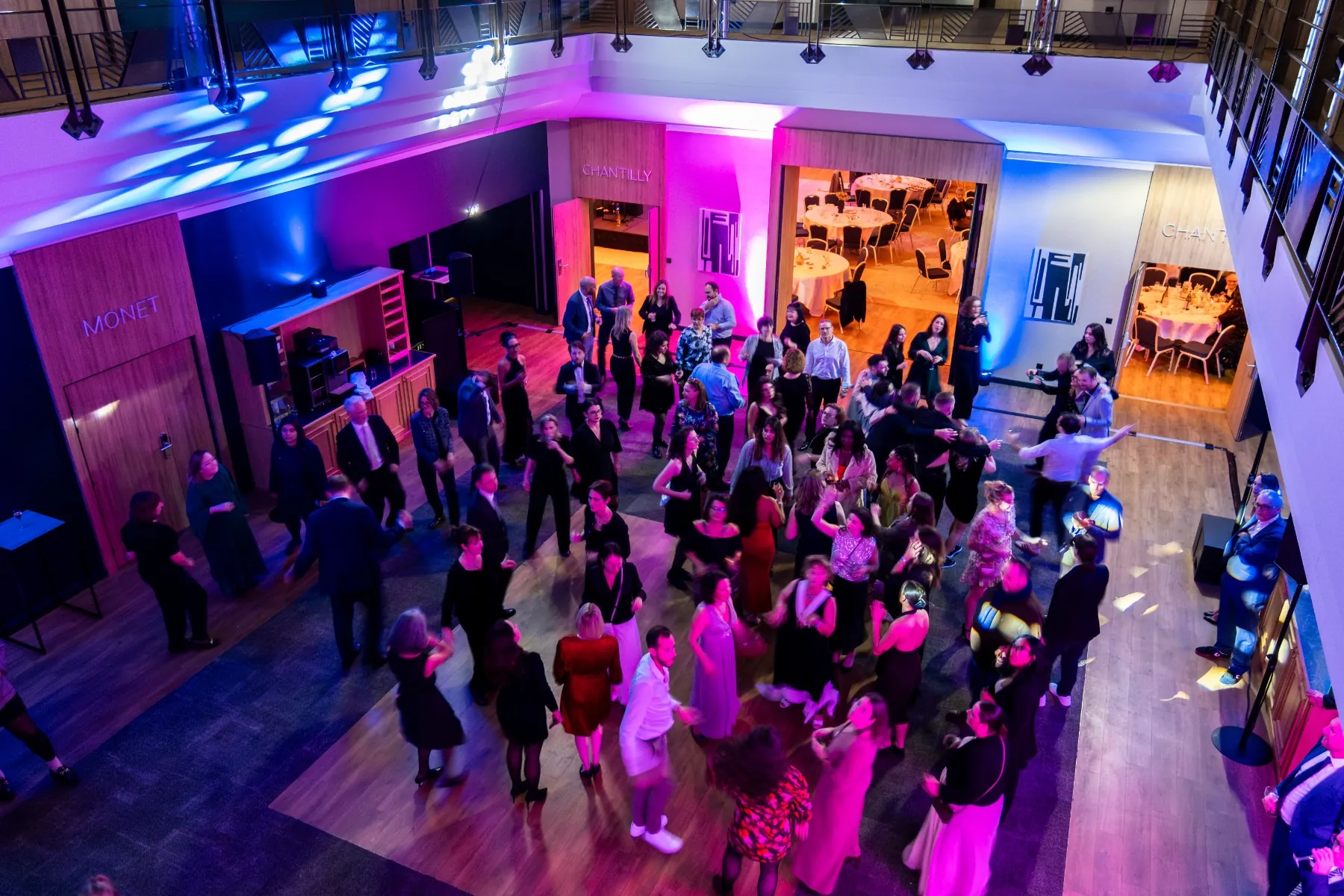 en::Photo of a dance party at the Mercure Chantilly, taken from the upper floor, showing festive lights, a DJ, guests dancing, and an adjoining room set up for dinner fr::Photo d’une soirée dansante au Mercure Chantilly, prise depuis l’étage, avec lumières festives, DJ, invités en train de danser et salle attenante dressée pour le dîner