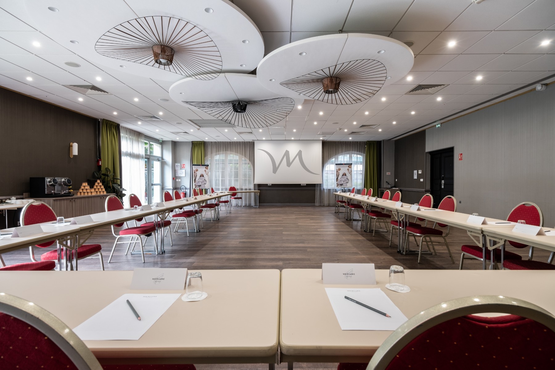 en:: a meeting room with red chairs and a projection screen, arranged in a large U-shape layout with a view of the hotel’s garden fr:: une salle de réunion avec des chaises rouges et un écran de projection, en disposition en U de grande capacité, avec vue sur le jardin de l’hôtel