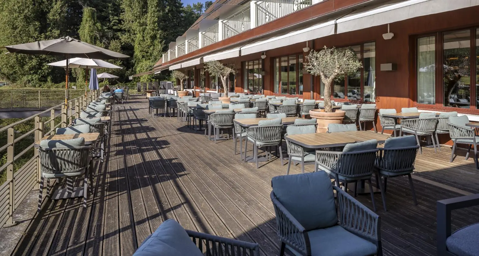 en::long view of the hotel terrace, with set tables, open parasols, cushions on chairs and olive trees, highlighting the seating capacity fr::vue en longueur de la terrasse de l’hôtel, avec tables dressées, parasols ouverts, coussins sur les chaises et oliviers, permettant d’apprécier la capacité d’accueil
