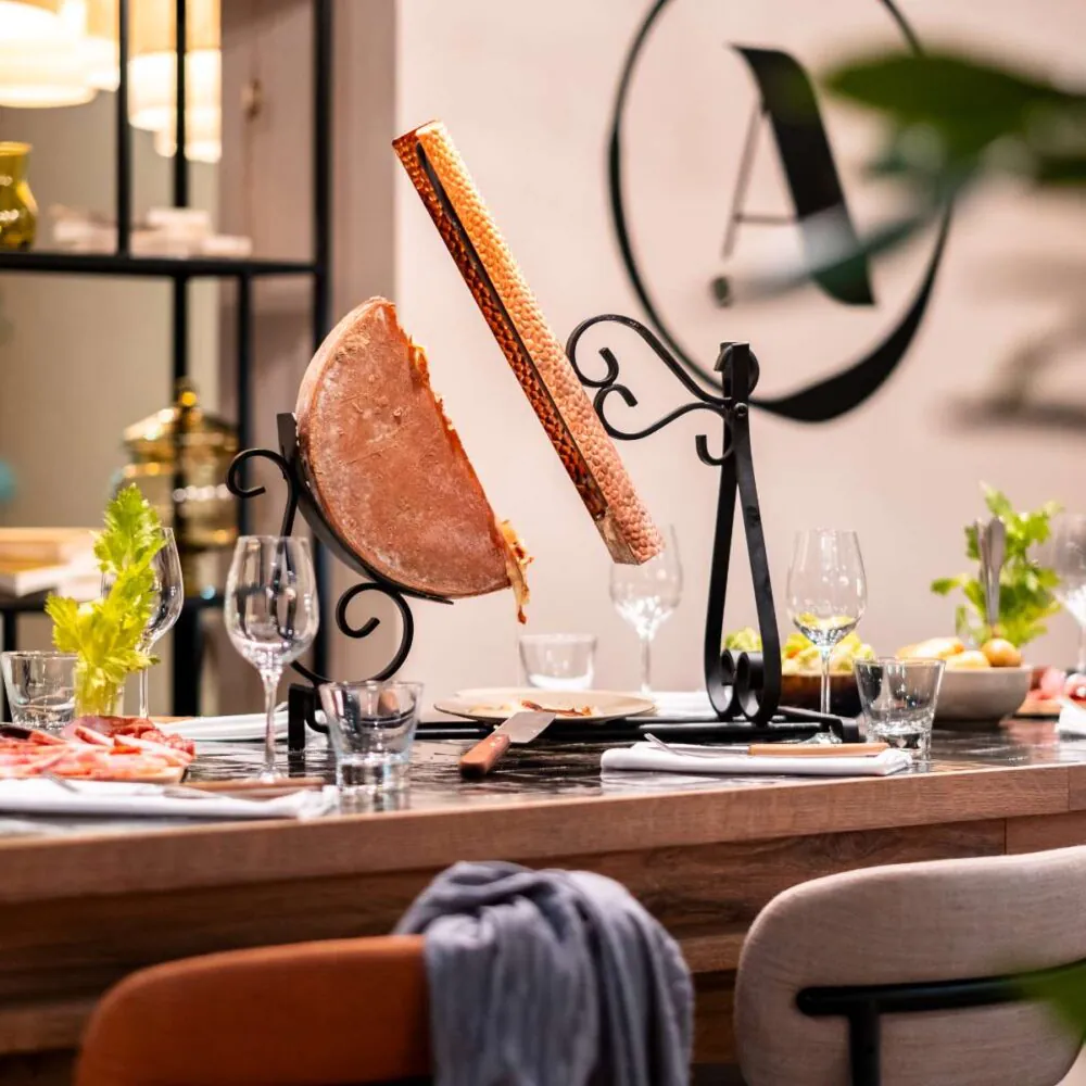 zoom sur une table haute en bois, dressée pour une raclette traditionnelle, avec pommes de terre, charcuterie et l’enseigne A l’Epicerie en arrière-plan