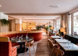 en::restaurant dining room viewed from the back of the space, with square tables for two or four set along the windows, round tables in alcoves with banquette seating, and a glimpse of the cheese dome, fine grocery display, buffet area and entrance corridor fr::salle de restaurant vue depuis un angle du fond, avec tables carrées de deux ou quatre le long des fenêtres, tables rondes en alcôves avec banquettes intégrées, aperçu de la cloche à fromages et du meuble d’épicerie fine, puis plus loin l’espace buffet et le couloir d’entrée