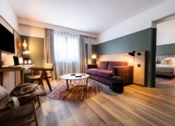 en::suite with a view of the lounge and work area in the foreground, featuring a two-seat mauve sofa, desk, chairs and wall-mounted TV, and the bedroom in the background fr::suite avec vue sur le salon et l’espace bureau au premier plan (canapé deux places mauve, bureau, chaises et TV murale), et la chambre en second plan, aux tons chauds, avec rideaux épais, voilages et parquet au sol
