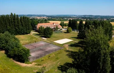 en::drone view of the hotel showing the main building, parking area, tennis and volleyball courts, pétanque area, and the surrounding forested park fr::vue de drone de l’hôtel montrant le bâtiment principal, le parking, les terrains de tennis et de volley, le boulodrome et le parc arboré entouré de forêt
