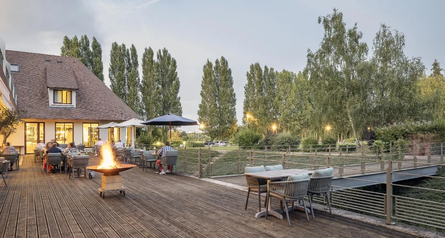 en::side view of the hotel’s ground-floor terrace, showing the outlook toward the park, with set tables, a lit fire pit and guests enjoying the space fr::vue latérale de la terrasse de l’hôtel, offrant un aperçu de la vue sur le parc, avec tables dressées, braséro allumé et personnes en train de profiter du lieu
