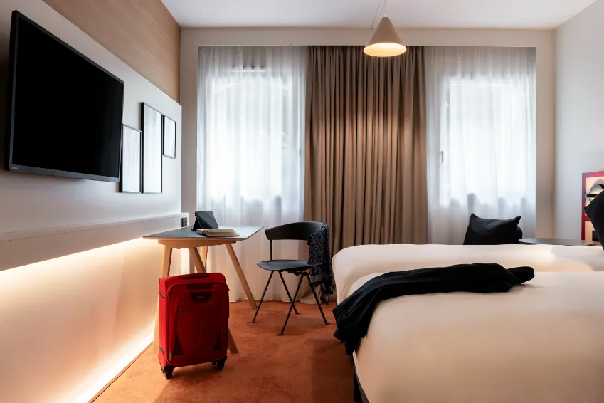 room with a metal-and-wood desk, warm lighting, twin beds, a red suitcase by the desk, and a coat on the bed