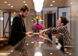 comptoir de réception avec l’accueil d’un client par une réceptionniste, et en arrière-plan la bibliothèque normande de l’hôtel