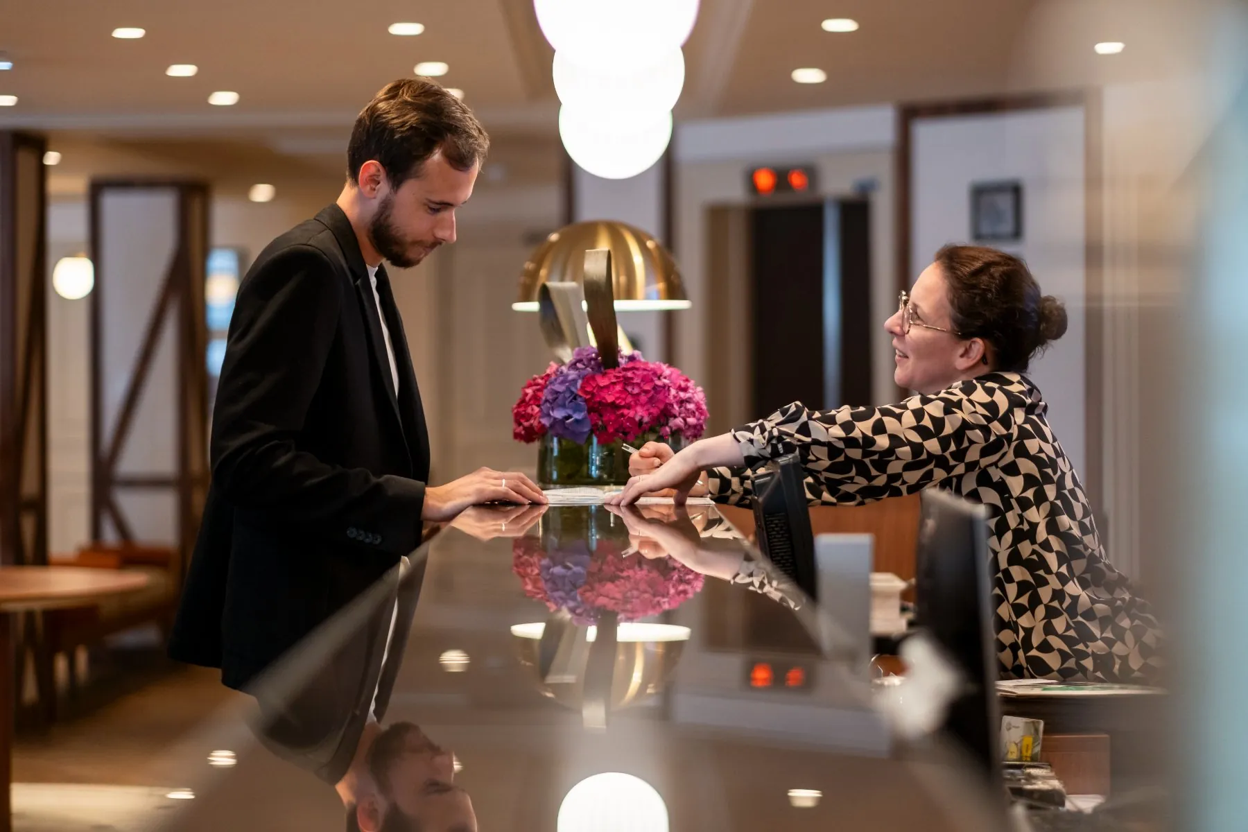 hotel reception counter with a guest being welcomed by a receptionist, with the hotel’s Norman-style library in the background