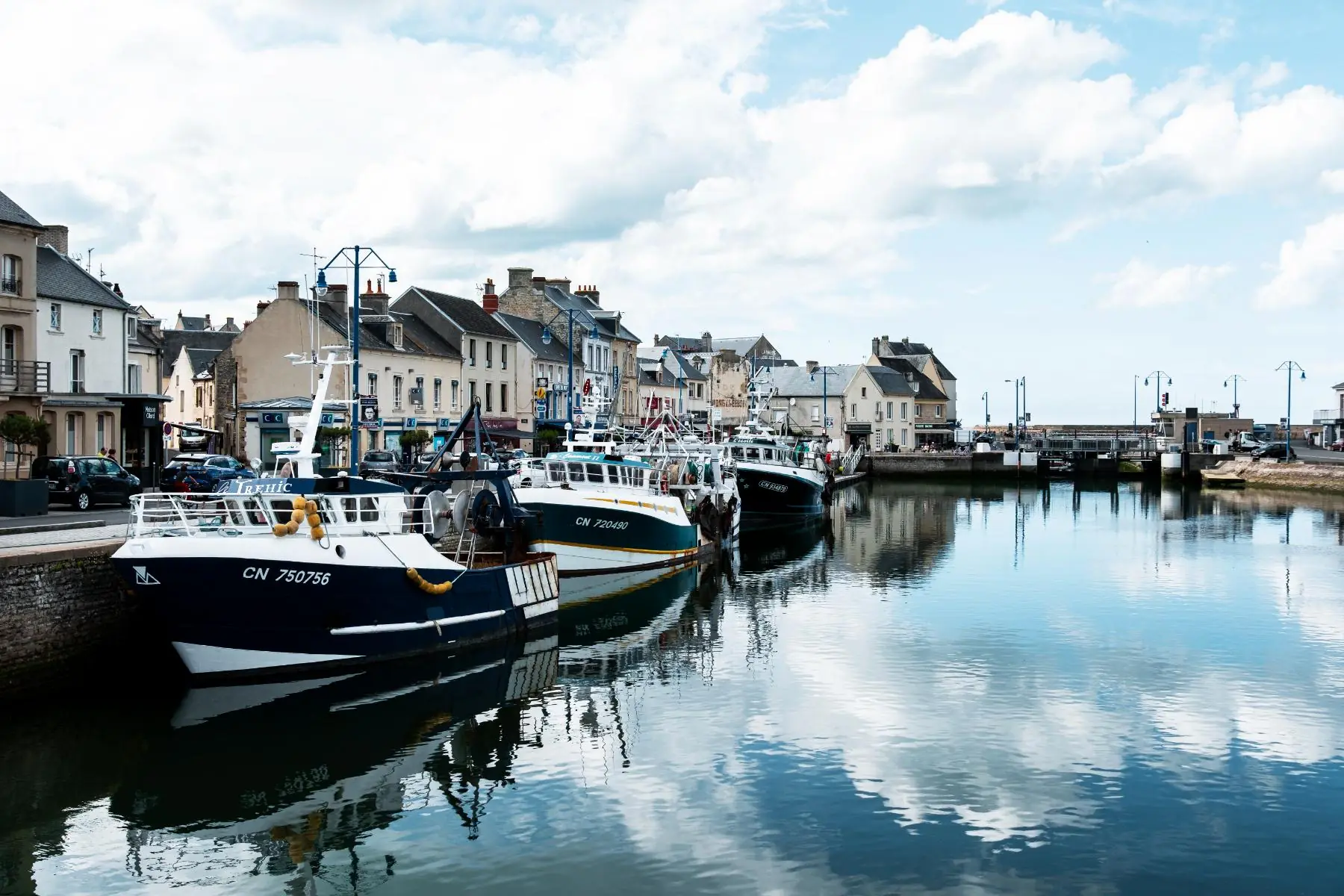 en::View of the port of Port-en-Bessin, with moored boats, cloud reflections on the water, and Norman houses and buildings in the background. fr::Vue du port de Port-en-Bessin, avec les bateaux amarrés, le reflet des nuages sur l’eau, et en arrière-plan des maisons et bâtiments normands.