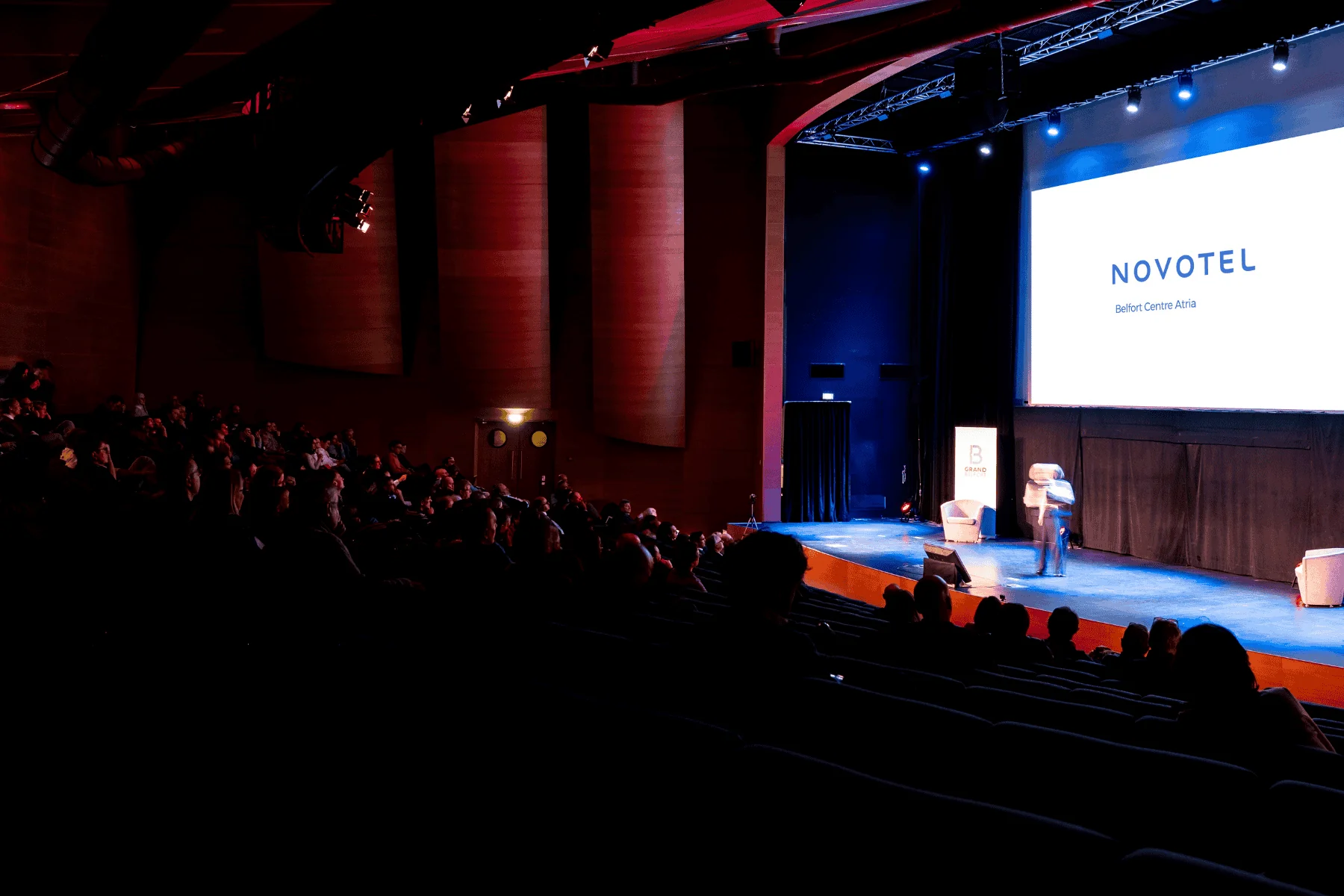 large auditorium with rows of seats, a Novotel Belfort Centre Atria screen, and a speaker addressing the audience