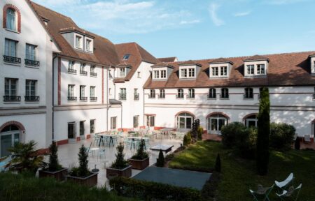 en::a view of the façade facing the inner gardens, with buildings forming an L-shape in the background and a furnished terrace in the foreground fr::vue de la façade côté jardins intérieurs, avec en arrière-plan des bâtiments formant un L et, au premier plan, une terrasse aménagée de tables et chaises