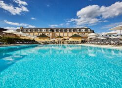 vue prise depuis la piscine extérieure en direction du bâtiment de l’hôtel, parasols et transats délimitant les espaces, avec le golf en arrière-plan