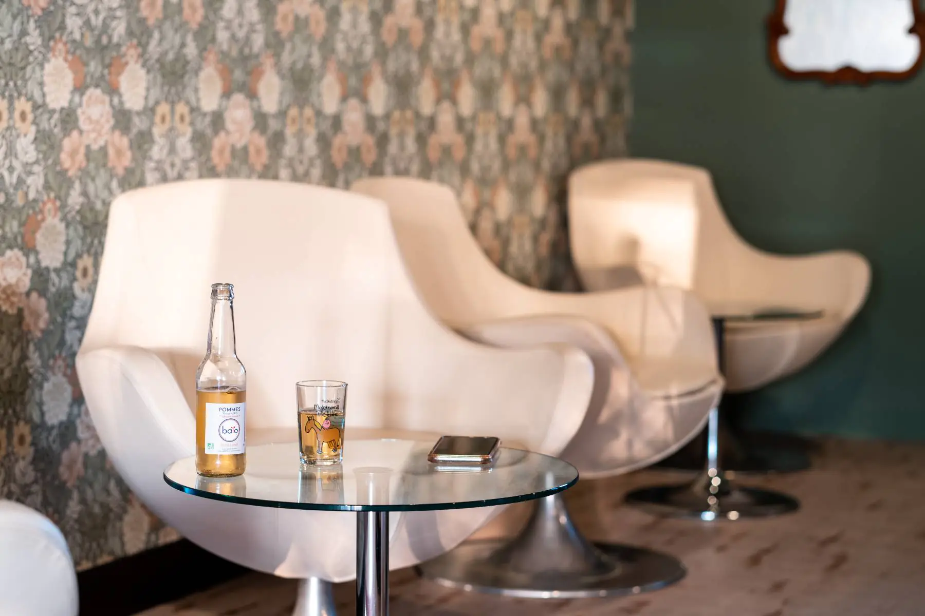 en::Seventies-inspired armchairs arranged around round transparent tables, one of which features an organic apple juice served in a playfully designed glass. fr::Fauteuils esprit années 70 disposés autour de tables rondes et transparentes, dont l’une accueille un jus de pomme bio servi dans un verre au design ludique.