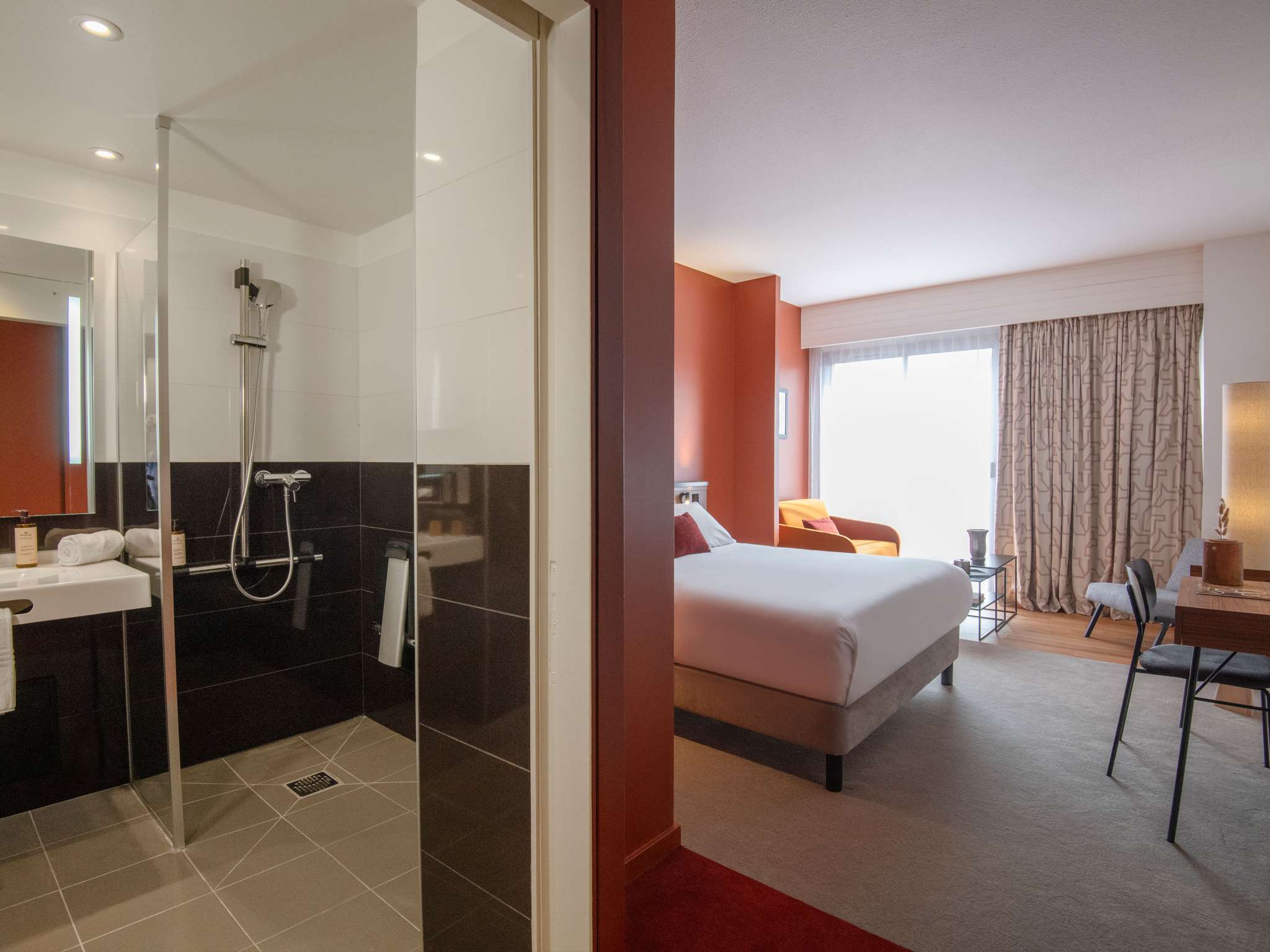 Vue depuis la chambre PMR montrant la salle de douche et l’espace nuit, avec lit double bordé d’une couette blanche et coussins rouges, fauteuil jaune orangé, fauteuil gris, bureau en bois, rideaux occultants beiges aux motifs géométriques et luminaires jaune orangé