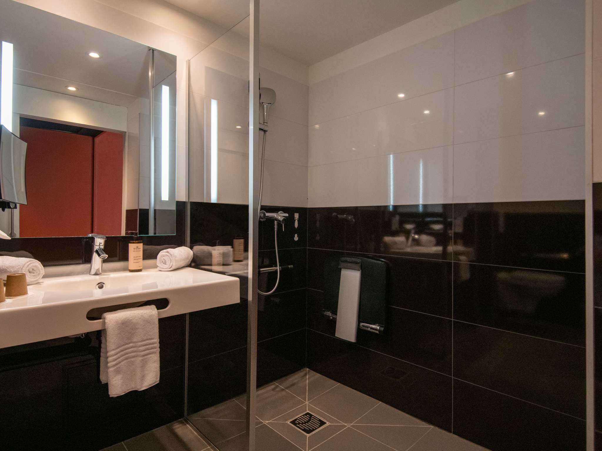 Salle de douche PMR avec lavabo blanc, grand carrelage au sol, faïence murale bicolore blanc et gris foncé, miroir rétroéclairé laissant apparaître le rouge de la porte de chambre