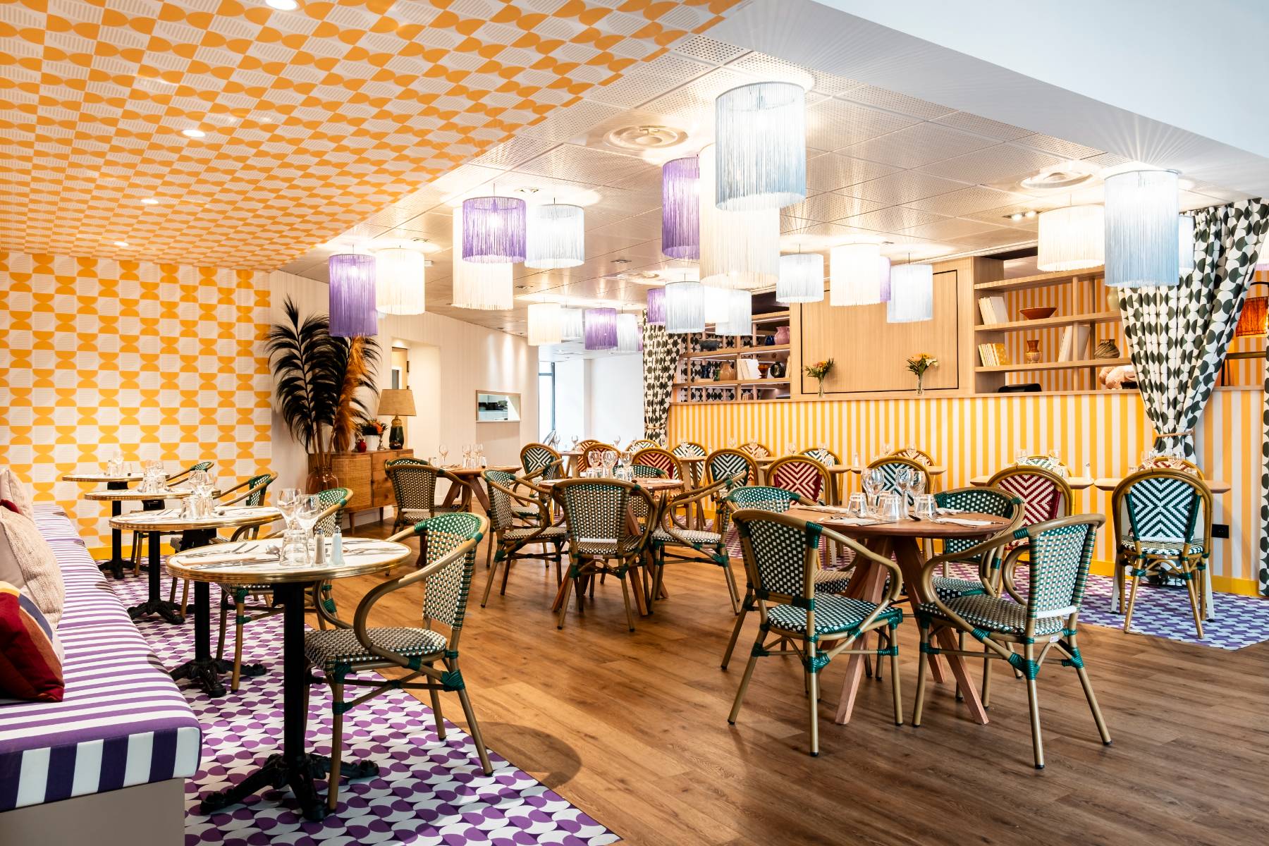 Salle de restaurant avec parquet au sol, murs et partie du sol ornés de motifs diabolo jaune et blanc ou mauve et blanc, luminaires à franges mauve, blanc et bleu gris, banquettes rayées mauve et blanc, tables bistrot, chaises en osier vert et blanc ou mauve et blanc, étagères décoratives et console en osier
