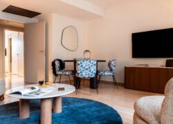 en::View of a lounge featuring, in the background, a wooden table with four blue-patterned fabric chairs, a designer mirror on the wall, in the foreground a coffee table and comfortable beige armchairs, and between a wooden storage unit with a wall-mounted television, creating a warm and homely atmosphere. fr::Vue sur un salon avec, en arrière-plan, une table en bois et quatre chaises en tissu bleu sur fond blanc, miroir design au mur, au premier plan table basse et fauteuils beiges confortables, et entre les deux : un meuble de rangement en bois avec télévision murale, créant une atmosphère chaleureuse et conviviale.