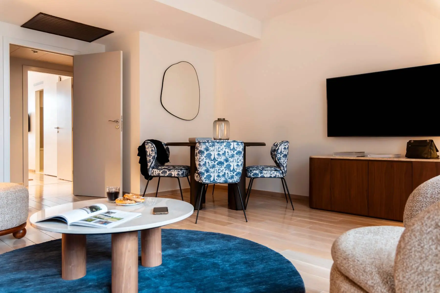 en::View of a lounge featuring, in the background, a wooden table with four blue-patterned fabric chairs, a designer mirror on the wall, in the foreground a coffee table and comfortable beige armchairs, and between a wooden storage unit with a wall-mounted television, creating a warm and homely atmosphere. fr::Vue sur un salon avec, en arrière-plan, une table en bois et quatre chaises en tissu bleu sur fond blanc, miroir design au mur, au premier plan table basse et fauteuils beiges confortables, et entre les deux : un meuble de rangement en bois avec télévision murale, créant une atmosphère chaleureuse et conviviale.