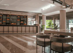 en::Bright hotel reception area accessible from the golf course, with bar and restaurant blinds in the background, beige and brown speckled tiled floor, black and beige graphic ceiling with hanging lights, almond-green front desk, black and orange shelving, and a high tab fr::Réception de l’hôtel lumineuse, accessible depuis le golf, avec stores du bar-restaurant en arrière-plan, sol carrelé beige et marron moucheté, plafond graphique noir et beige avec luminaires suspendus, desk vert amande, étagères noires et orangées, et table haute avec chaises de bar pour patienter ou travailler.