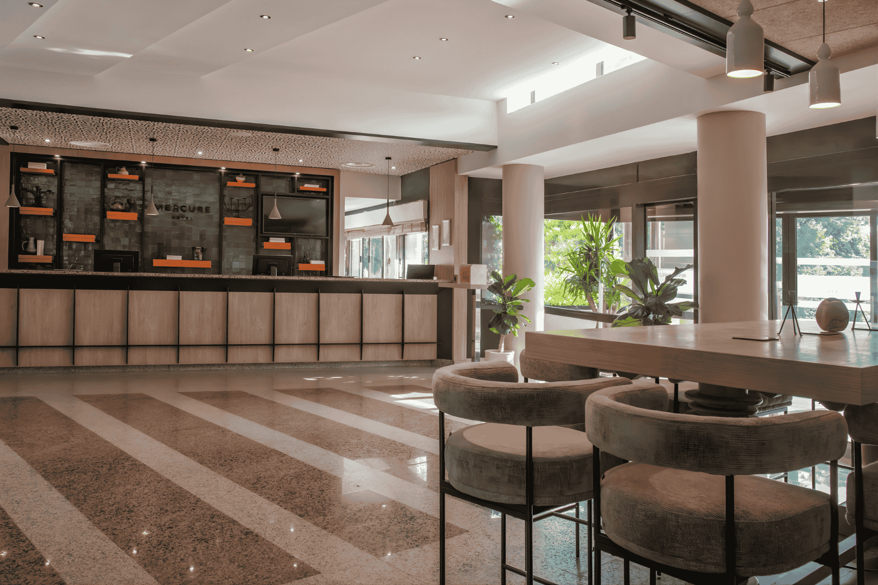 en::Bright hotel reception area accessible from the golf course, with bar and restaurant blinds in the background, beige and brown speckled tiled floor, black and beige graphic ceiling with hanging lights, almond-green front desk, black and orange shelving, and a high tab fr::Réception de l’hôtel lumineuse, accessible depuis le golf, avec stores du bar-restaurant en arrière-plan, sol carrelé beige et marron moucheté, plafond graphique noir et beige avec luminaires suspendus, desk vert amande, étagères noires et orangées, et table haute avec chaises de bar pour patienter ou travailler.