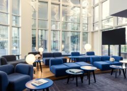 en::Glass-walled lobby area with views over Paris Boulogne, furnished with blue sofas and armchairs, blue and black coffee tables, an integrated screen, and round table lamps. fr::Espace lobby vitré avec vue sur Paris Boulogne, aménagé de canapés et fauteuils bleus, de tables basses bleues et noires, d’un écran intégré et de luminaires ronds posés sur les tables.