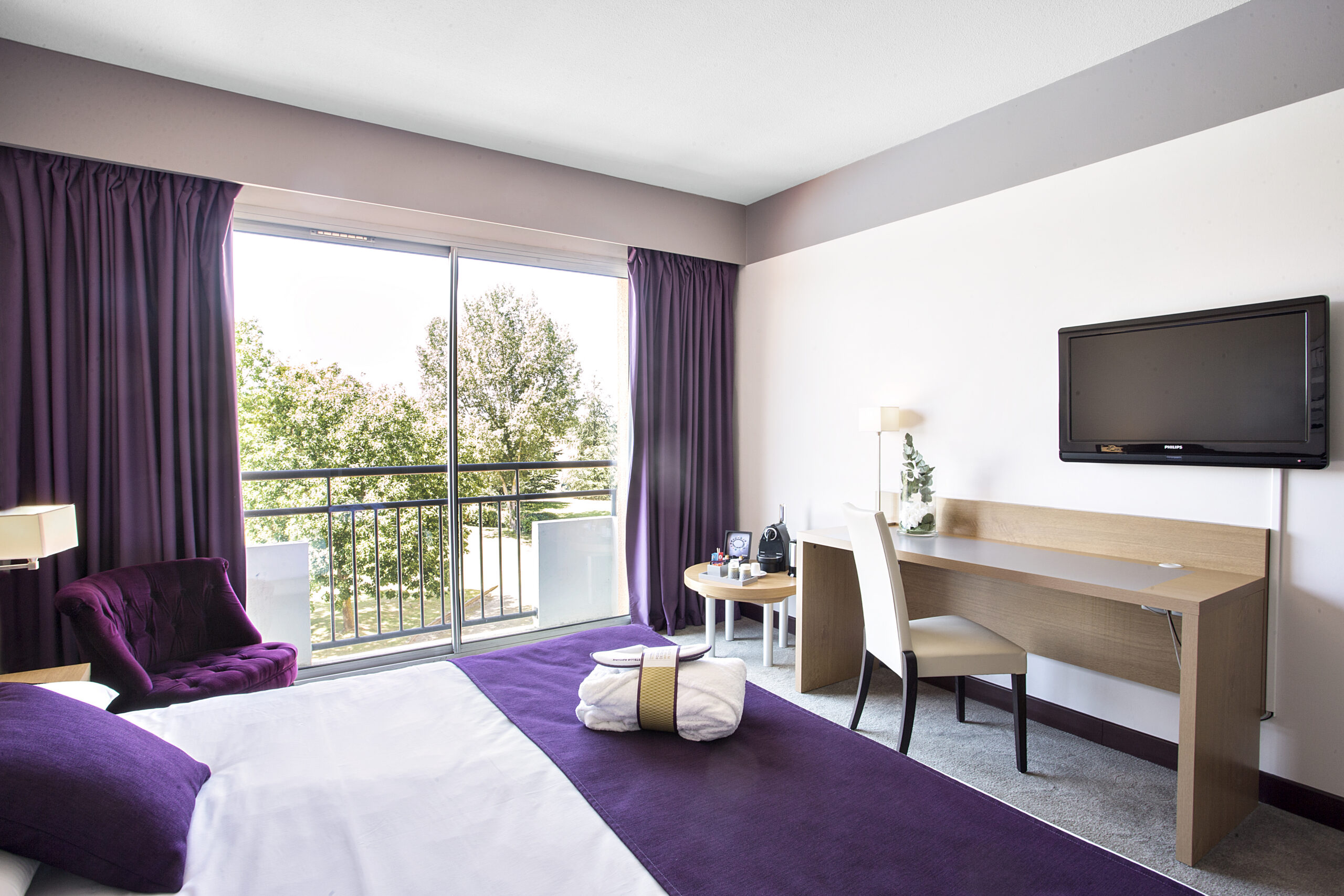 Chambre avec vue sur le golf, lit double avec couvre-lit et coussins mauves, fauteuil mauve et bureau en bois clair en face avec télévision murale et machine Nespresso