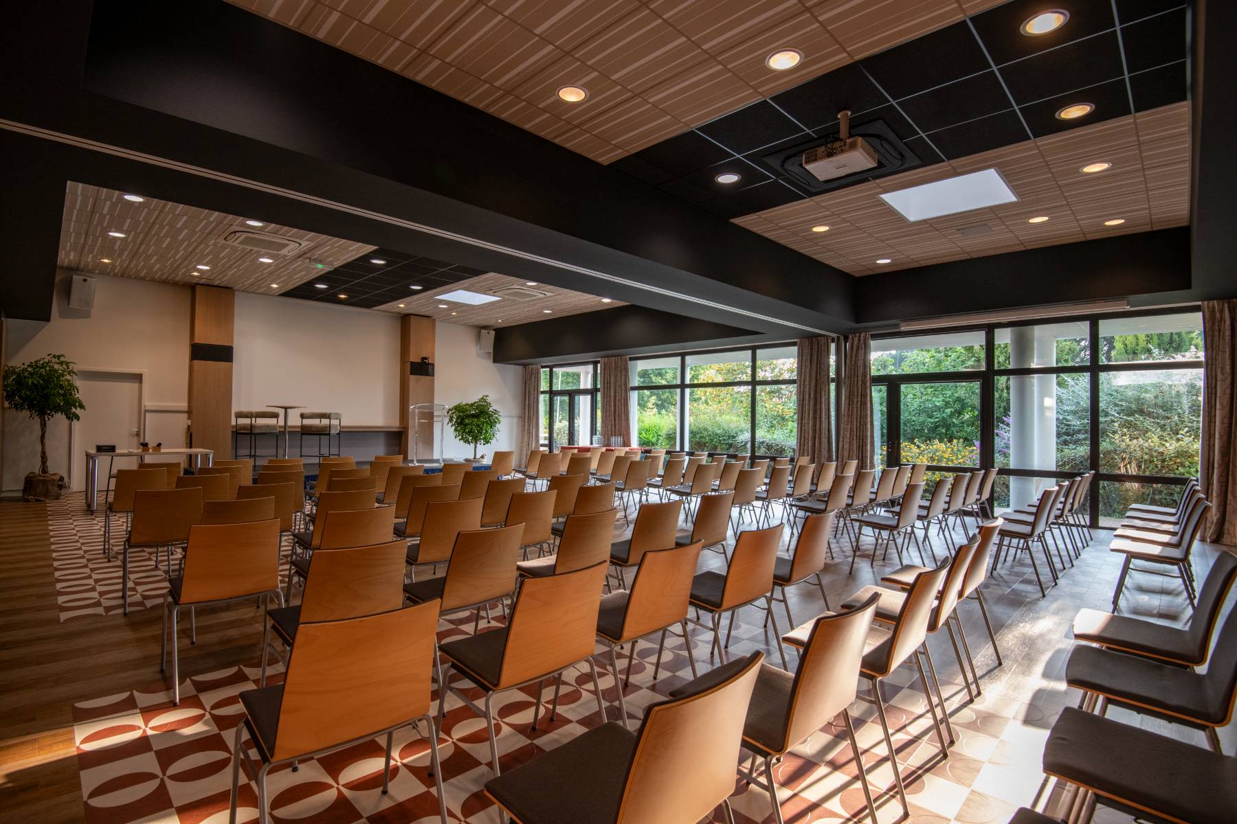 Rows of chairs are lined up in a bright room with large windows overlooking greenery and a wall-mounted screen