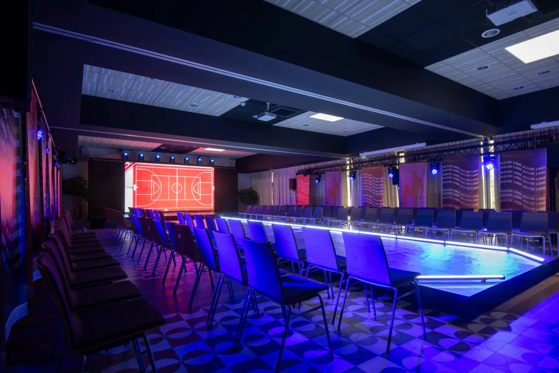 Dark room with rows of chairs facing a lit stage and a red and white screen