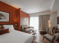 en::Suite with red walls inspired by Toulouse, featuring a double bed with red cushions, a two-seater grey sofa bed with red cushions, a coffee table, ottoman, lounge chair, console, and wall-mounted television defining the living area. fr::Suite aux murs rouges rappelant la couleur de Toulouse, avec lit double et coussins rouges, canapé-lit gris à deux places avec coussins rouges, table basse, pouf, fauteuil de détente, console et télévision murale délimitant l’espace salon.