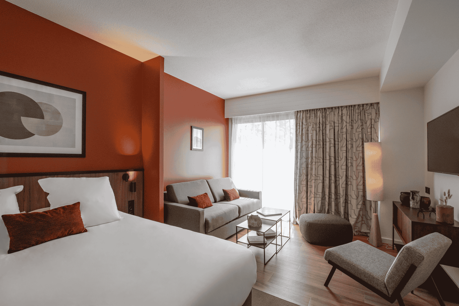 en::Suite with red walls inspired by Toulouse, featuring a double bed with red cushions, a two-seater grey sofa bed with red cushions, a coffee table, ottoman, lounge chair, console, and wall-mounted television defining the living area. fr::Suite aux murs rouges rappelant la couleur de Toulouse, avec lit double et coussins rouges, canapé-lit gris à deux places avec coussins rouges, table basse, pouf, fauteuil de détente, console et télévision murale délimitant l’espace salon.