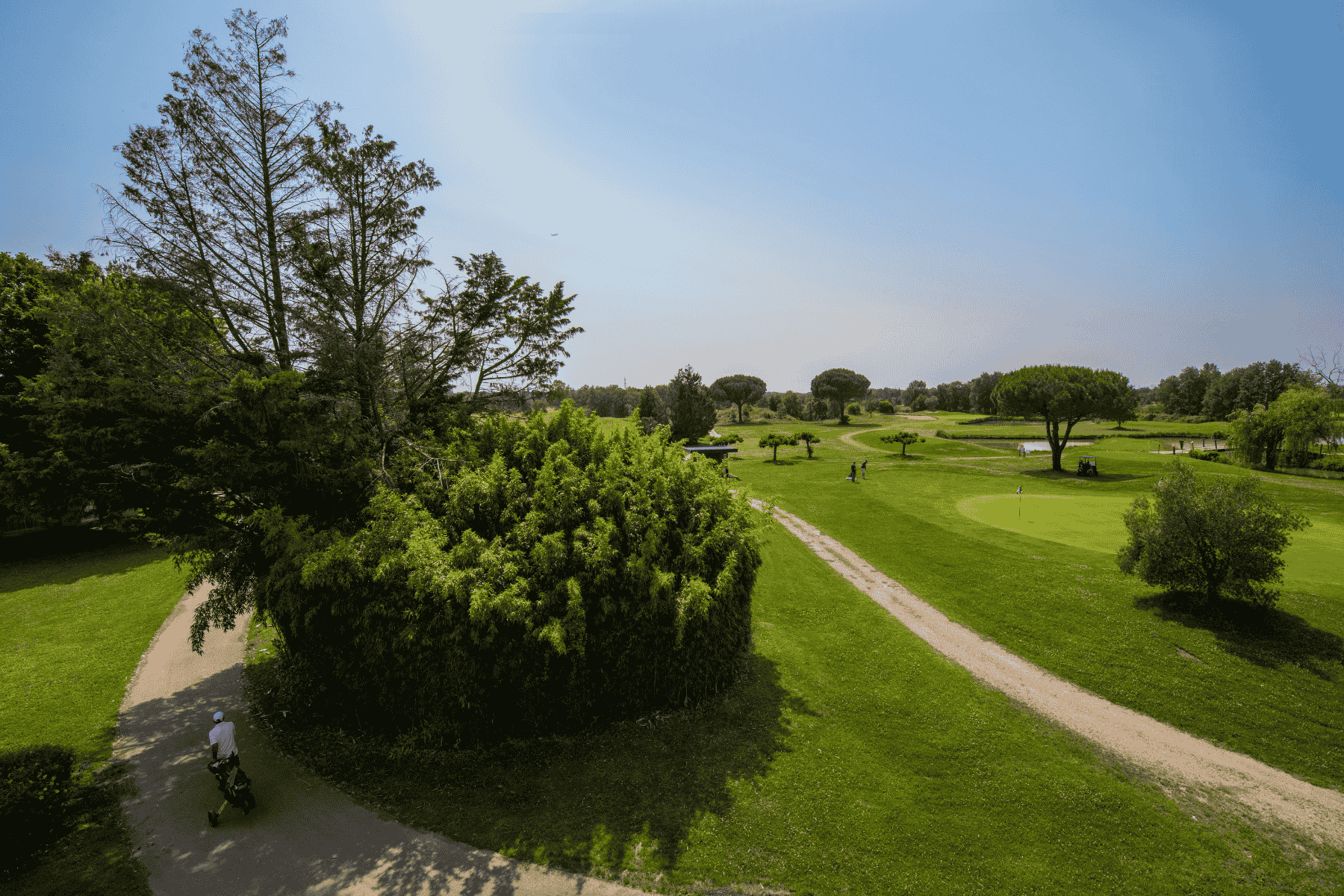 en::View from a hotel room over the 18-hole golf course, with a tree-lined path, a golfer seen from behind pulling a trolley, a nearby green with two players, water features, and lush vegetation. fr::Vue depuis une chambre sur le golf 18 trous, avec un chemin bordé d’arbres, un golfeur de dos tirant son chariot, un green proche avec deux joueurs, des points d’eau et une végétation abondante.