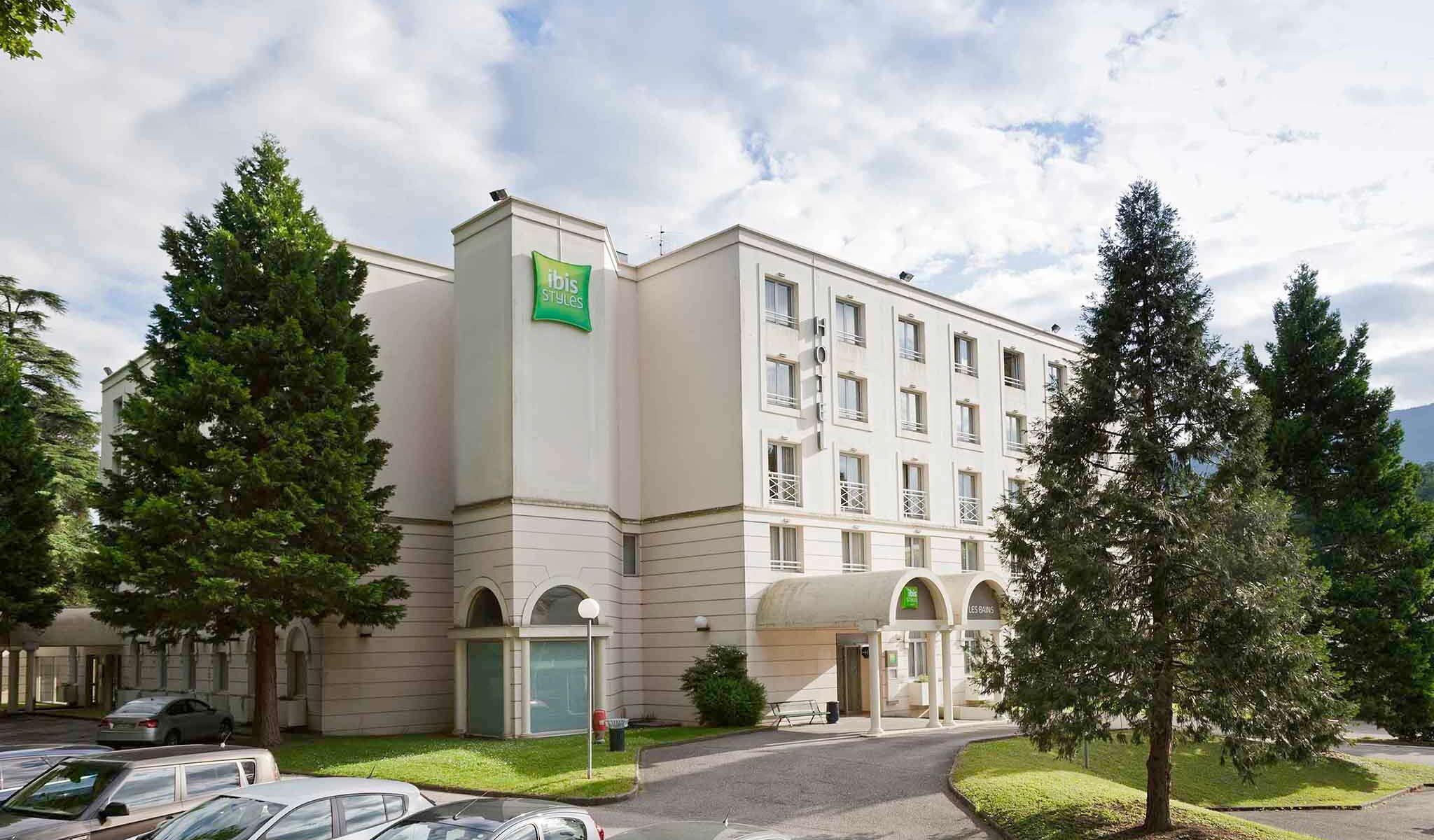 en::White building façade with green ibis Styles sign, parked cars, pine tree and lawn. fr::Façade de bâtiment blanc avec enseigne ibis Styles verte, voitures stationnées, sapin et pelouse.