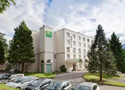 en::White building façade with green ibis Styles sign, parked cars, pine tree and lawn. fr::Façade de bâtiment blanc avec enseigne ibis Styles verte, voitures stationnées, sapin et pelouse.