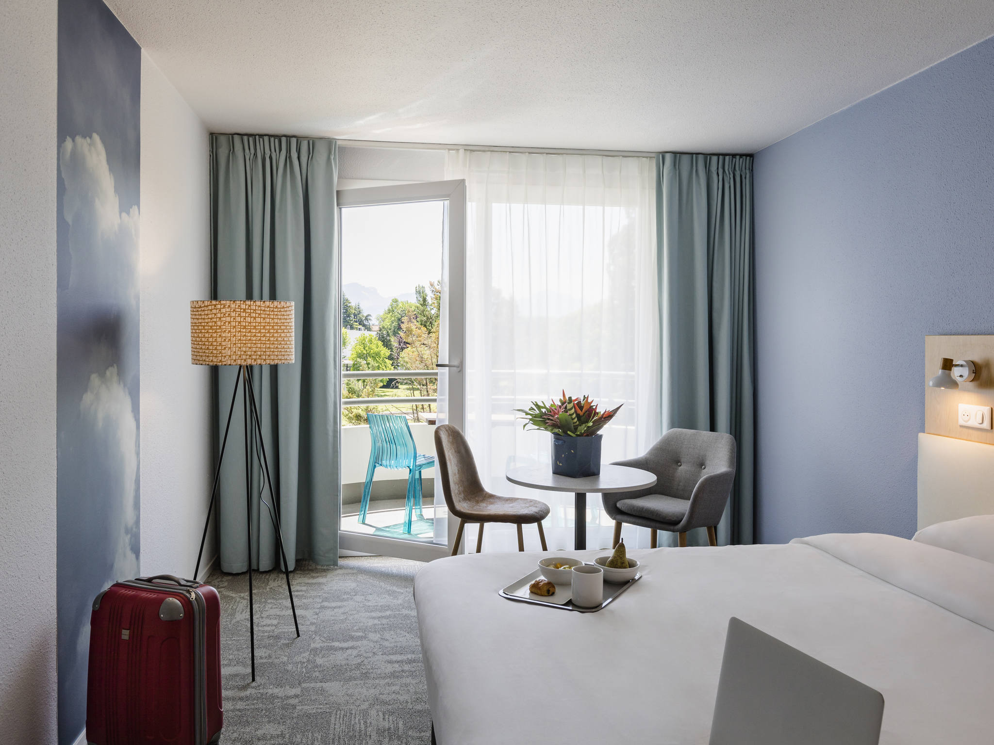 en::Double bed with breakfast tray, red suitcase near the wall, bouquet of flowers on a table near the balcony access with turquoise blue chair through a large glass door. fr::Lit double avec plateau petit-déjeuner, valise rouge près du mur, bouquet de fleurs sur une table près de l’accès au balcon avec chaise bleu turquoise par une grande porte-fenêtre.