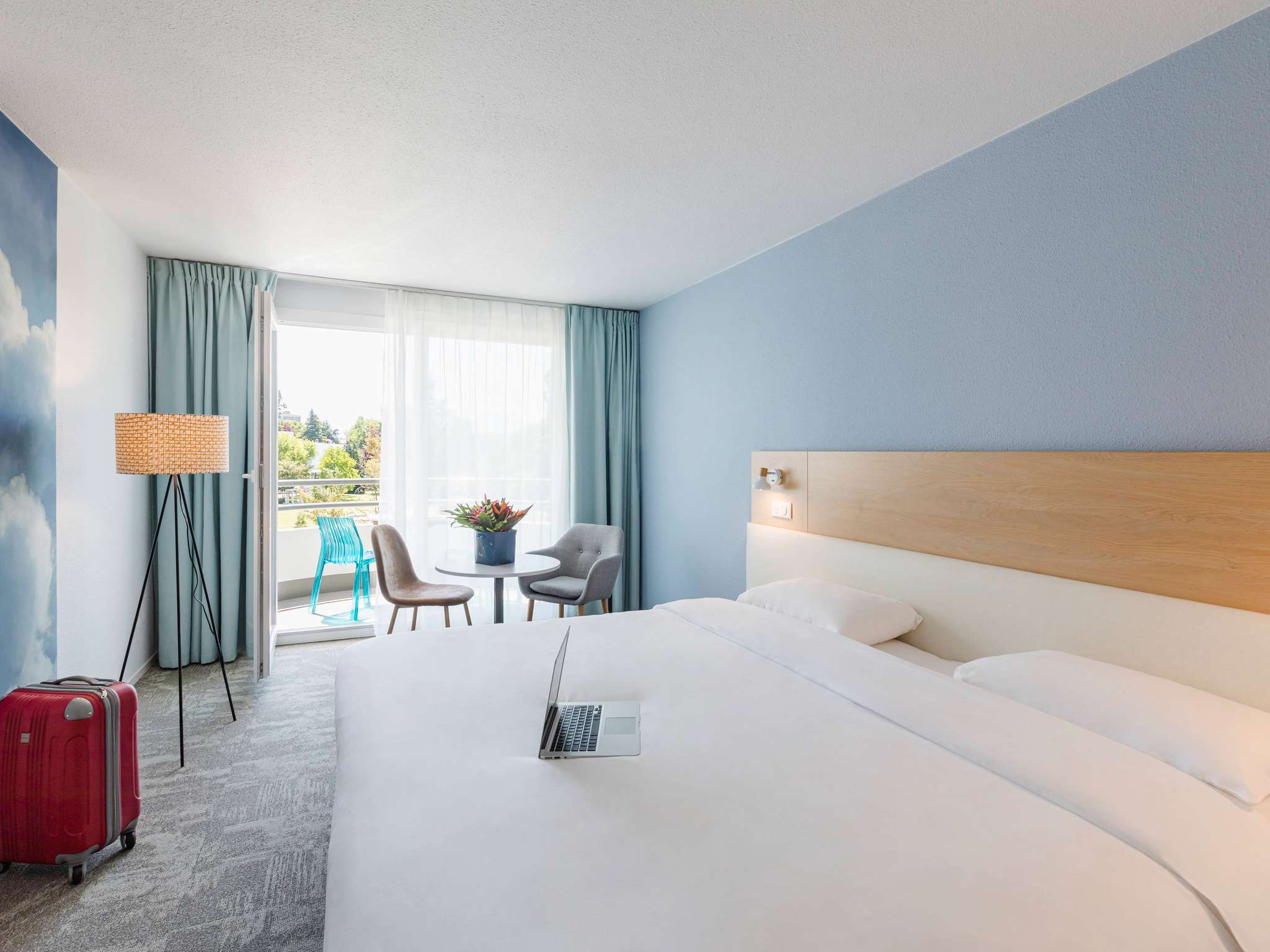 en::Double bed with light wood headboard and white finish, large wall photo of blue sky with clouds, bouquet of flowers on a table near the balcony access through a large glass door. fr::Lit double avec tête de lit en bois clair et revêtement blanc, grande photographie de ciel bleu avec nuages au centre du mur, bouquet de fleurs sur une table près de l’accès au balcon par une grande porte-fenêtre.