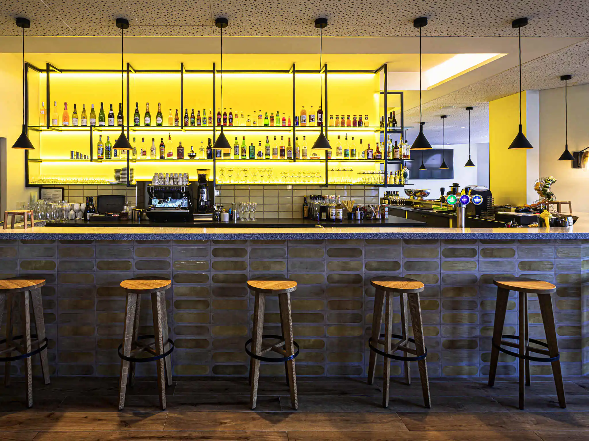 en::Tiled bar counter in brown tones with marble finish, wooden stools with round seats, backlit drinks wall in bright yellow.fr::Comptoir bar carrelé dans les tons bruns avec finition marbre, tabourets en bois, mur de boissons rétroéclairé jaune.