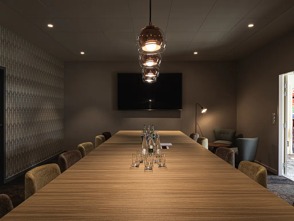 en::Long wooden table with glasses and glass water bottles, round pendant lights above, wall-mounted TV facing. fr::Grande table en bois en longueur avec verres et bouteilles d’eau en verre, luminaires ronds suspendus, écran TV mural en face.