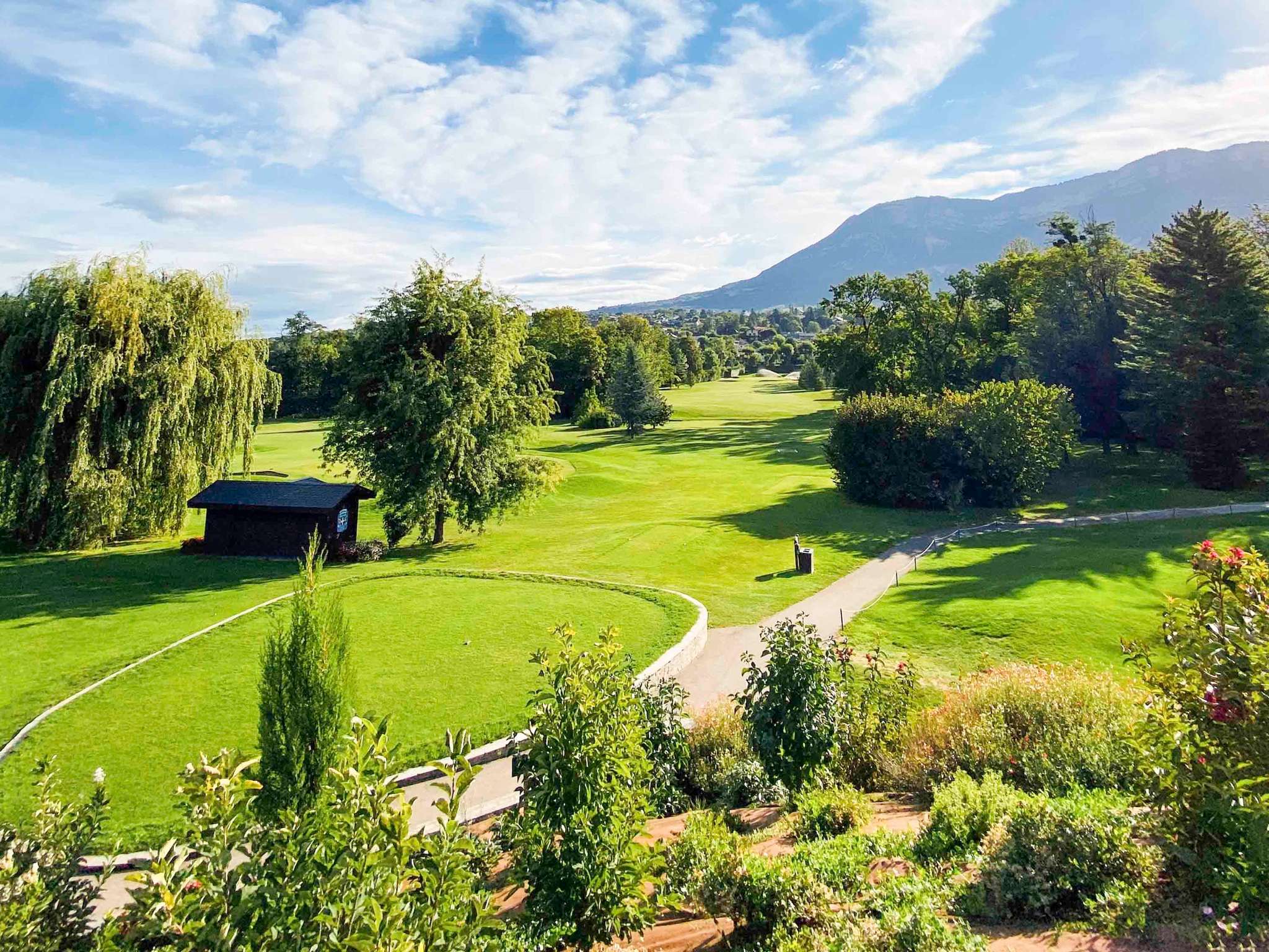en::Park with trees and pines, alpine mountains in the background, wooden cabin between two trees. fr::Parc avec arbres et sapins, monts alpins en arrière-plan, cabane en bois entre deux arbres.