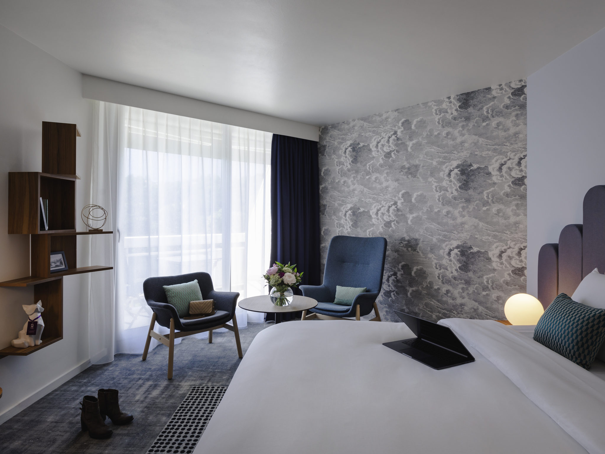 en::Double bed with an open laptop, wooden shelves with a fox figurine, books and photo frame, chairs around a round coffee table. fr::Lit double avec ordinateur ouvert, étagères en bois avec renard décoratif, livres et cadre photo, chaises autour d’une table basse ronde.