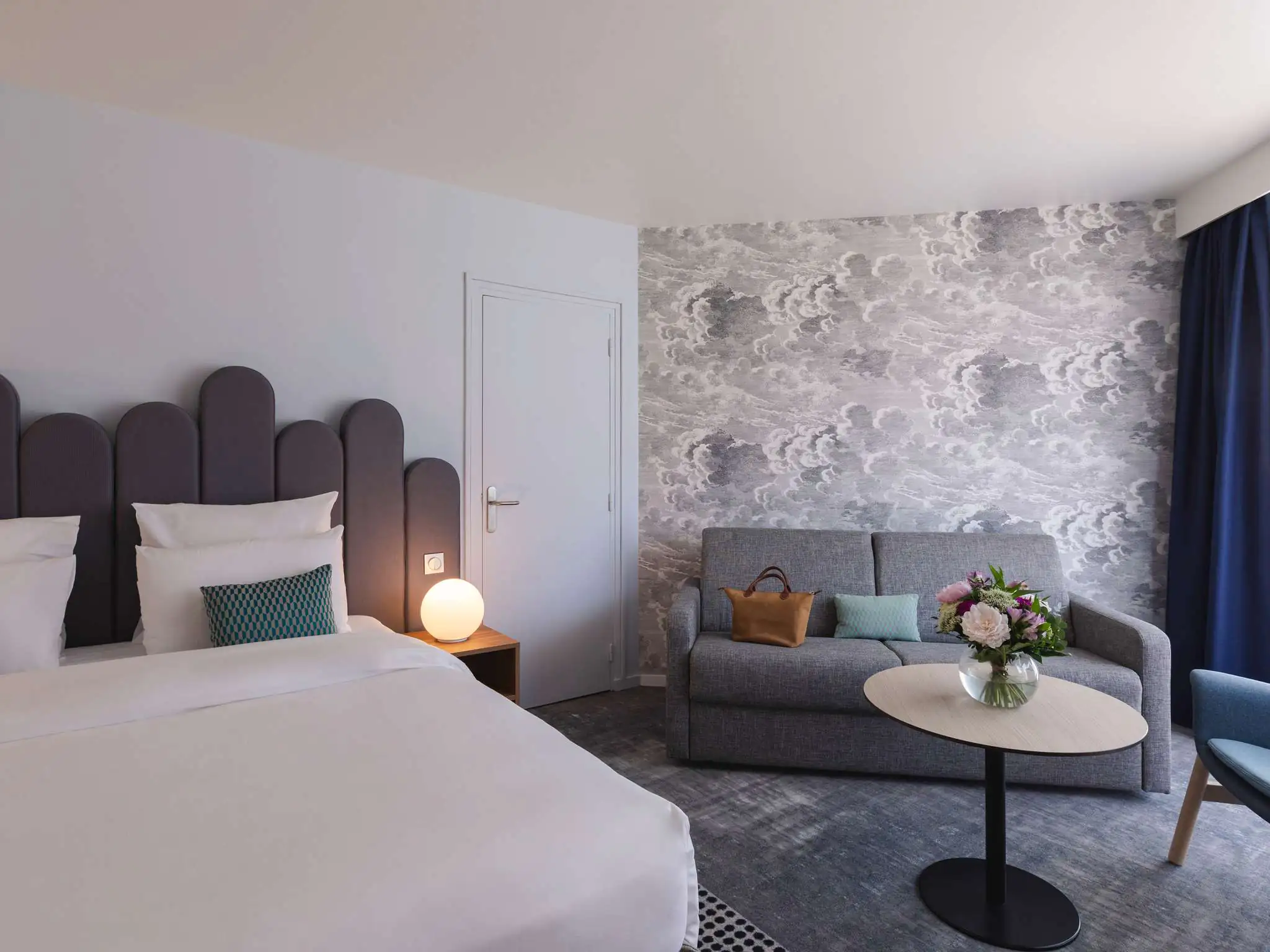 en::Double bed with a grey headboard, double sofa in front of a wall covered in grey cloud-patterned wallpaper, coffee table with a bouquet of flowers. fr::lit double avec tete de lit grise, canapé double devant un mur tapissé de nuages gris table basse avec bouquet de fleurs