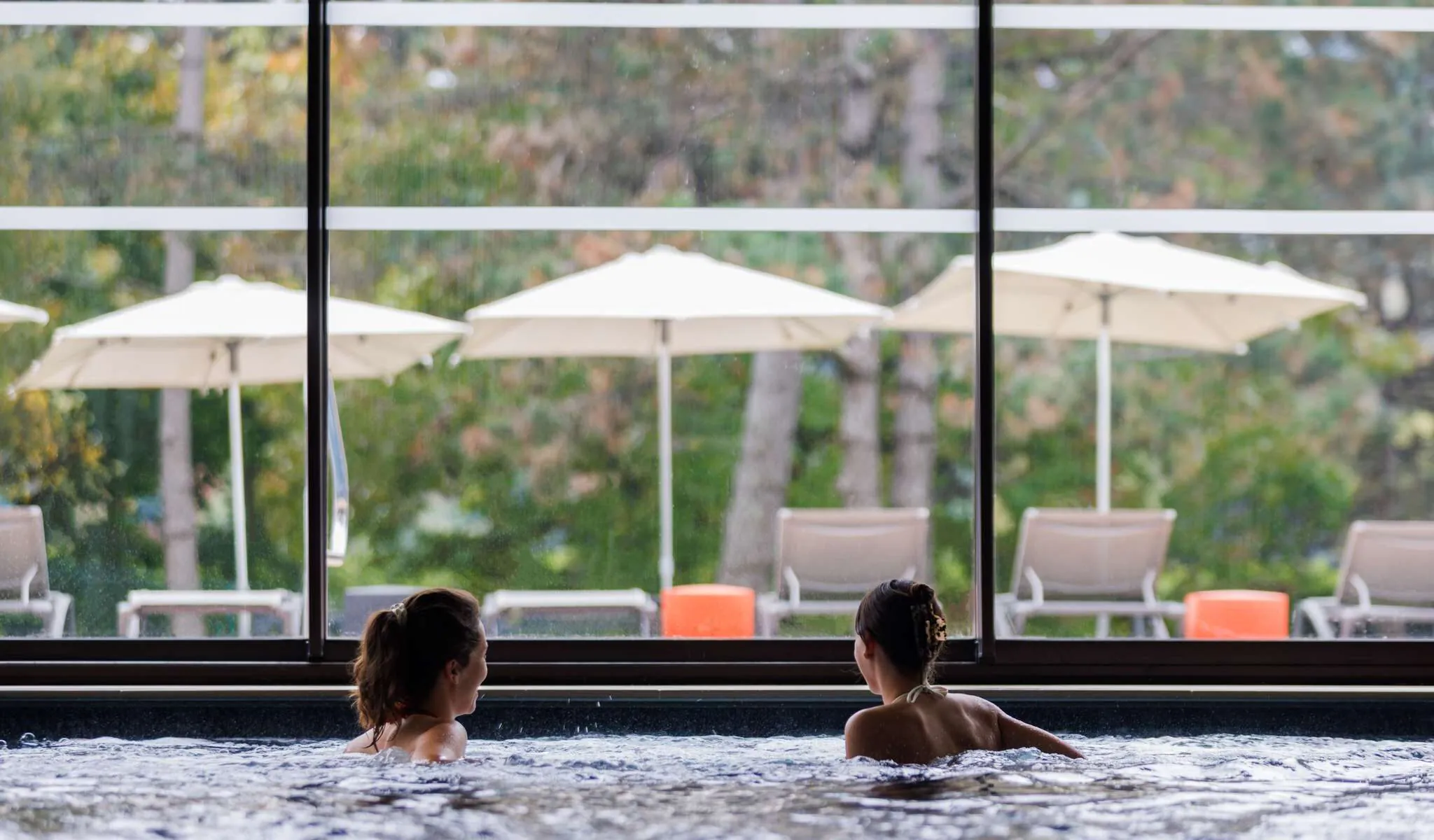 en::Pool with hot tub, two people relaxing with their backs turned, facing a bay window overlooking the park and solarium. fr::Piscine avec bain à remous, deux personnes de dos se détendant face à une baie vitrée ouvrant sur le parc et le solarium.
